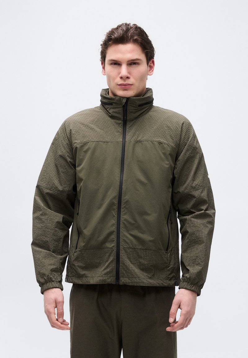 Νεαρός άνδρας που φοράει μπουφάν με κουκούλα σε ελαιοπράσινο χρώμα, με φερμουάρ μπροστά και υφή στα μανίκια και τους ώμους, στέκεται μπροστά σε απλό φόντο.