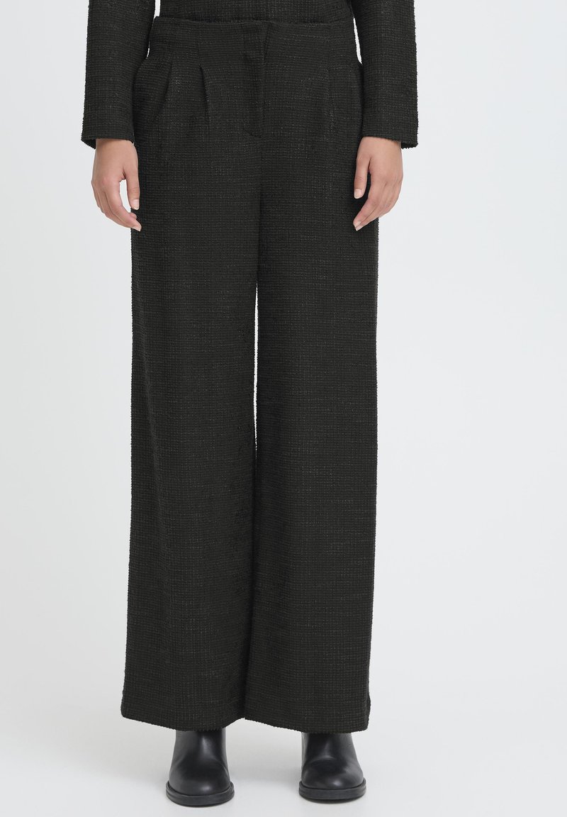 Pantaloni neri a gamba larga realizzati con tessuto strutturato, caratterizzati da pieghe frontali e tasche laterali, abbinati a stivali neri alla caviglia.