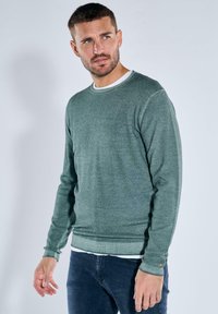 Grüner Strickpullover mit rundem Ausschnitt, gerippten B�ündchen und Saum, über einem weißen Hemd getragen, kombiniert mit dunklen Jeans. Glatte Textur.