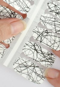Nagelfolien verfügen über einen glänzenden weißen Grund mit filigranen schwarzen Wirbelmustern. Sie werden zur Anwendung auf einem silbernen Untergrund gehalten.