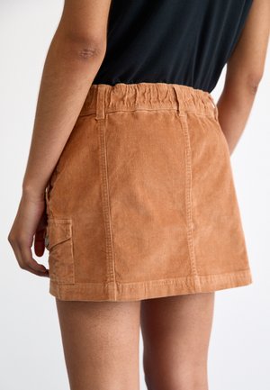 Kvinde iført en beige kort buksekjole i kordfløjl med elastisk talje og sidelomme, kombineret med en sort top.
