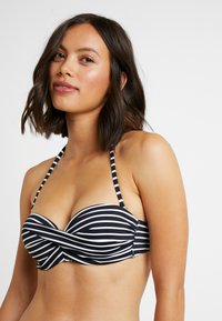 Bikini vršek s černobílým pruhovaným vzorem a zkrouceným designem vpředu, s tenkými ramínky a bandeau střihem. Hladká textura.