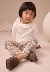 Biały top z falbankowym kołnierzem, beżowe legginsy w kwiatowy wzór oraz brązowe zamszowe buty. Dziecko siedzi na jasnym podłożu.