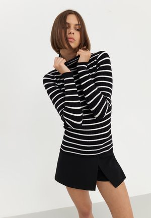 Svart og hvit stripete ribbet turtleneck-genser med lange ermer, sammen med svarte shorts med splitt på siden. Enkel, tettsittende design.