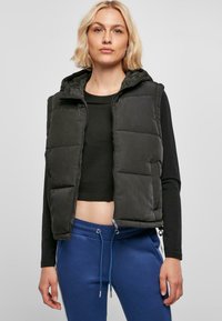 Crni softness prsluk s kapuljačom, prednjim zipom i bočnim džepovima, nosi se preko crnog kratkog topa i plavih dukserica s detaljima uzice.