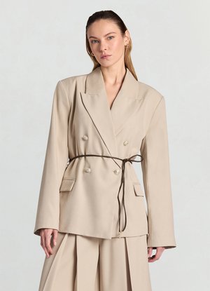 Blazer beige à double boutonnage avec larges revers, manches longues et taille ceinturée. Comprend deux poches avant et de grands boutons décoratifs.