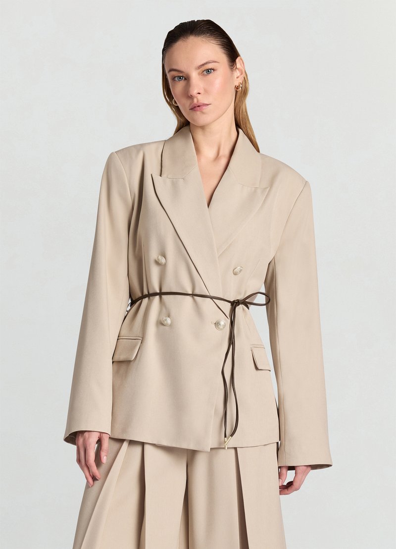 Blazer beige à double boutonnage avec larges revers, manches longues et taille ceinturée. Comprend deux poches avant et de grands boutons décoratifs.