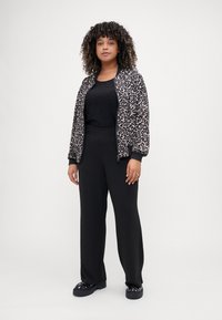 Jachetă bomber cu imprimeu de leopard, cu manșete negre din tricot, asortată cu un top negru strălucitor și pantaloni negri lejeri. Prezintă încălțăminte masivă neagră.