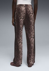 Πίσω όψη ατόμου που φοράει φαρδιά παντελόνια με καφέ λεοπάρ στροπή και μαύρες ρίγες στο πλάι, με καφέ παπούτσια σε γκρι φόντο.