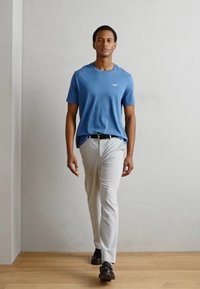 Blauwe katoenen t-shirt met korte mouwen en een wit logo, gecombineerd met witte broeken en zwarte schoenen, staand op een houten vloer.