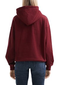 Hoodie croppato bordeaux con un grande cappuccio, vestibilità ampia e spalle basse; texture liscia con polsini a coste; abbinato a jeans scuri.