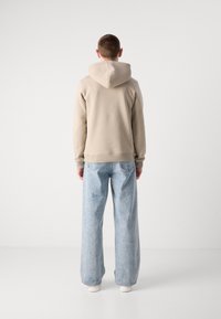 Bež kapuca s vrvico in rebrastimi manšetami, v kombinaciji s svetlo modrimi širokimi jeans hlačami. Materiali se zdijo mehki in sproščeni.