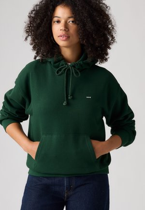 Donna con capelli ricci che indossa una felpa verde scuro con cappuccio e tasca frontale, e jeans blu, mani nella tasca della felpa, sfondo neutro.
