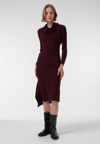 Bordeaux ribbel gebreide jurk met een colhalslijn en asymmetrische zoom, gecombineerd met kniehoog zwarte laarzen. Gladde textuur.