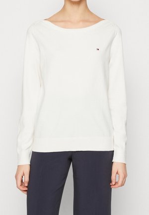 Biały sweter z długim rękawem, dekoltem w łódkę i małym logo Tommy Hilfiger na piersi, w połączeniu z ciemnymi spodniami.