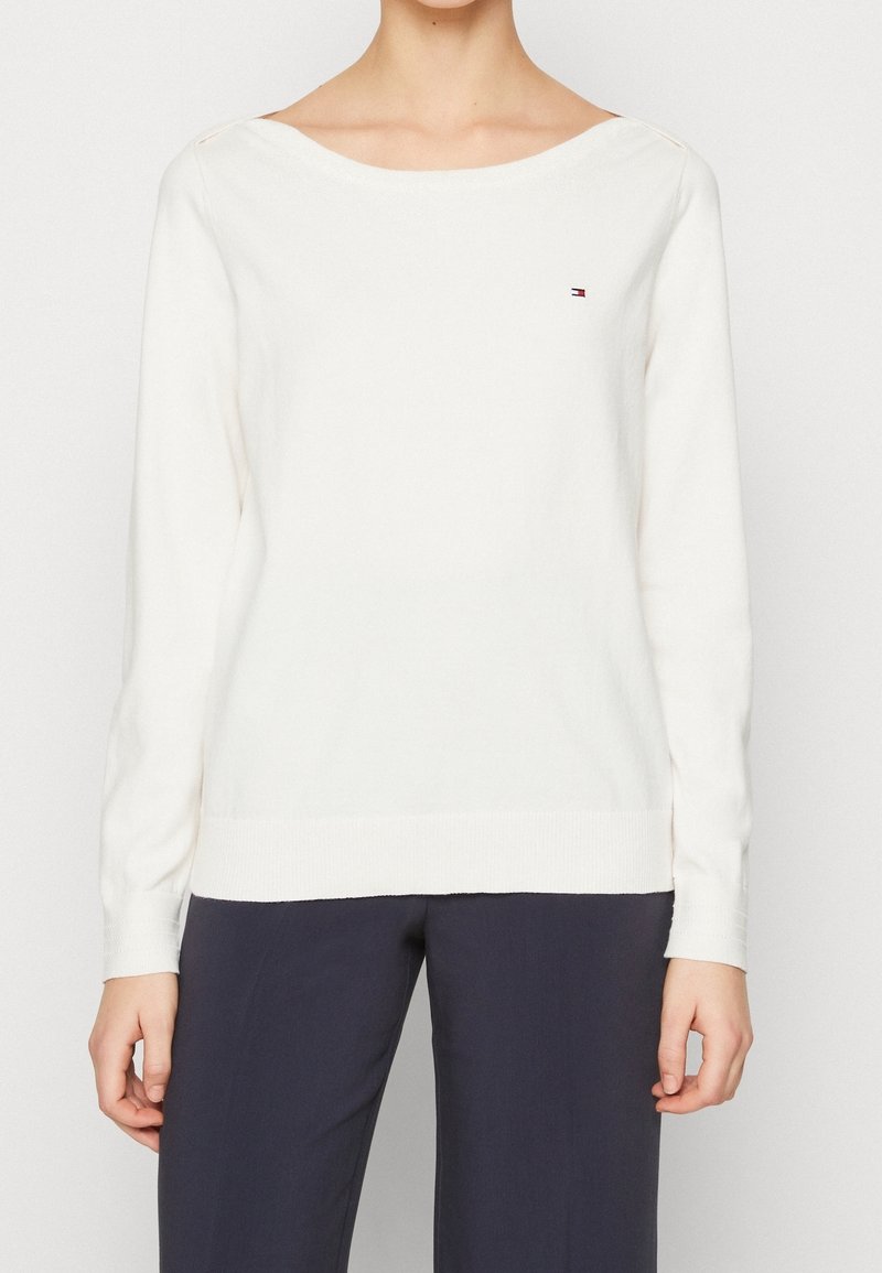 Pull blanc à manches longues avec encolure bateau et petit logo Tommy Hilfiger sur la poitrine, associé à un pantalon foncé.