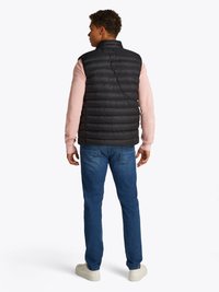 Colete puff preto com costura vertical, usado sobre uma camisa de manga longa rosa claro. Combinado com jeans azuis e ténis brancos.