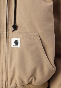 Tan zip-up jas gemaakt van stevig materiaal, met een zwarte trekkoord, voorkeeltas en een witte logo-patch met een zwart ontwerp.