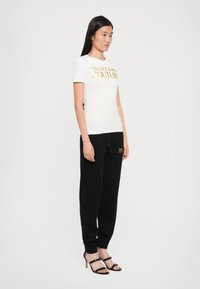 Camiseta blanca de manga corta con el logo dorado "Versace Jeans Couture", combinada con pantalones de chándal negros con detalles dorados. Sandalias de tacón negras.