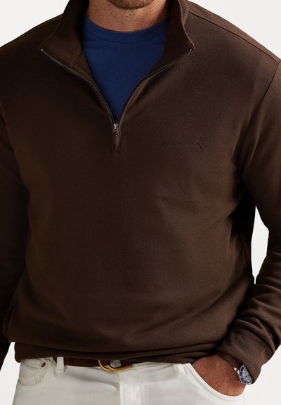 Bruine quarter-zip pullover in een gestructureerde stof, gedragen over een blauwe shirt, gecombineerd met witte broek en een horloge. Subtiele logo-detail op de borst.