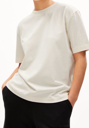 Personne portant un T-shirt à manches courtes couleur blanc cassé avec les mains dans les poches d'un pantalon noir, montrée du cou aux hanches.