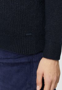 Gebreide trui in donker marineblauw met een gestructureerd patroon, ribgebreide manchetten en een klein zwart logo-label aan de onderkant. Gecombineerd met corduroy stof.