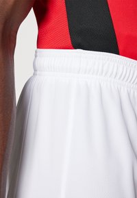 Witte sportshorts met een elastische tailleband, gemaakt van een gestructureerde stof. De top is rood met zwarte accenten en heeft een mesh-structuur.