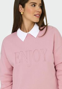 Vrouw in een roze sweatshirt met "ENJOY" reliëf, een witte blouse eronder en gouden gestructureerde ronde oorbellen, kijkend naar rechts.