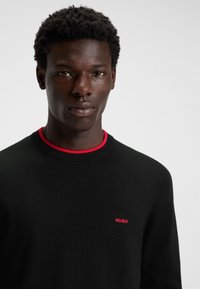 Schwarzer Strickpullover mit rundem rotem Kragen und kleinem rotem "HUGO"-Logo auf der linken Brust. Glatte Textur und figurbetonte Silhouette.