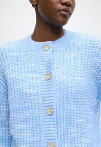 Ανοιχτό μπλε πλεκτή ζακέτα με στρογγυλό λαιμό, που διαθέτει τρία μεγάλα μπεζ κουμπιά, υφή υφάσματος και ριμπ λεπτομέρειες στο γιακά.