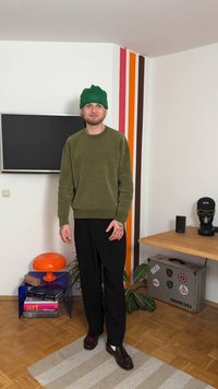 Groene gebreide muts, olijfgroene trui, zwarte broek met wijde pijpen en donkere schoenen. Binnenruimte met oranje lamp en patroon op de muur.