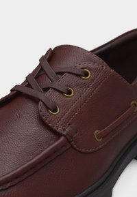 Pantof de barcă maro din piele cu șiret cu două găuri, ochiuri din alamă, cusături detaliate și talpă neagră din cauciuc vizibilă dedesubt.