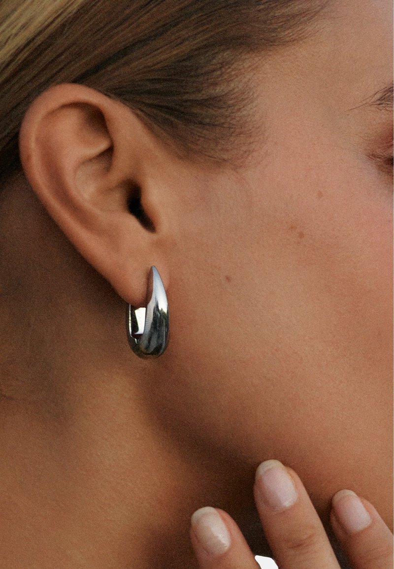 Leštený strieborný kruh so zaobleným tvarom a špicatým koncom. Hladký, reflexný povrch s moderným estetikou.