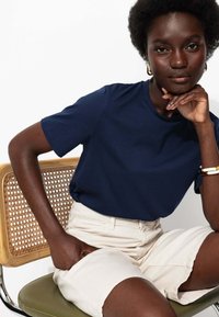 T-shirt en coton bleu marine avec un col rond, associé à un short beige. Modèle assis sur une chaise verte avec un dossier en osier, les mains posées.