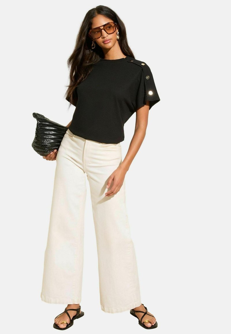 Schwarzes geripptes Kurzarmoberteil mit Knopfakzenten, weit geschnittene cremefarbene Hose und schwarze geflochtene Tasche. Schwarze Sandalen mit einer goldenen Akzentuierung runden den Look ab.