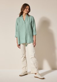 Camicia di lino verde chiaro con scollo a V e maniche a tre quarti, abbinata a jeans a zampa d'elefante off-white e sneakers beige.