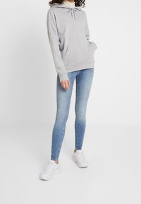 Mujer vestida con una sudadera gris claro, zapatillas blancas y jeans ajustados azul degradado, de pie contra un fondo blanco liso.