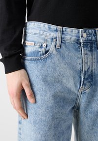 Light blue denim jeans with a faded pattern, five-pocket design, and a Calvin Klein label on the back waistband.