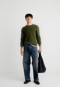 Jeune homme portant un pull vert olive et un jean bleu, tenant un sac fourre-tout noir, debout devant un fond blanc uni.