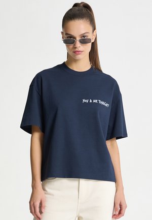 Giovane donna con i capelli lunghi raccolti in una coda di cavallo, che indossa piccoli occhiali da sole rettangolari, una maglietta blu navy con il testo "YOU & ME TONIGHT" e pantaloni bianchi.