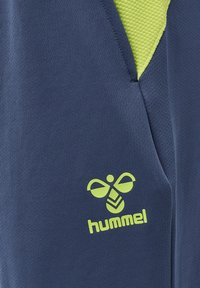Nærbillede af blåt stof med neongult Hummel-logo og matchende neongult mesh-lommefor.