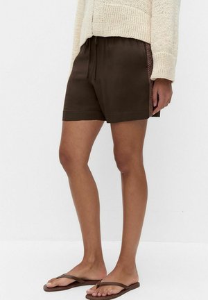 Personne portant un short marron foncé avec des panneaux en dentelle sur les côtés, un pull en maille beige et des tongs marron, debout sur un fond blanc uni.