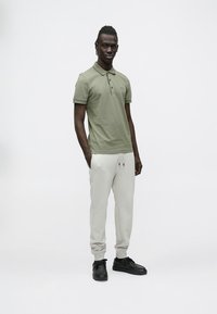 Zielona oliwkowa koszulka polo, jasnoszare spodnie dresowe ze sznurkiem, czarne trampki oraz stojąca sylwetka mężczyzny na białym tle.