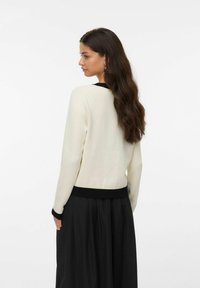 Ljust beige tröja med svarta ribbade fållar och muddar, med en enkel design och mjuk textur, bärs över en svart kjol.
