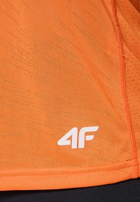 Pomarańczowa sportowa koszulka z teksturowaną tkaniną, ozdobiona ukośnymi paskami i białym logo w dolnym rogu. Gładki, lekki materiał.