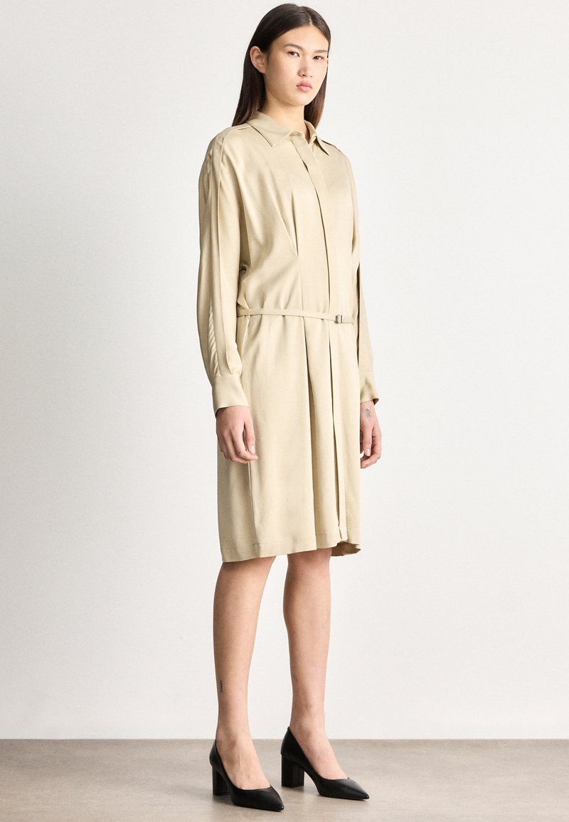 Mladá žena oblečená v béžových košeľových šatách so zúženým pásom a čiernych lodičkách na blokovom podpätku, stojaca pred jednoduchým svetlým pozadím.