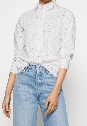 Vrouw draagt een wit, lang mouwen, knoophemd met zwarte stippen en strepen, ingestopt in lichtblauwe high-waisted jeans.