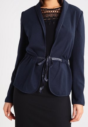 Vrouw draagt een marineblauw open voorjasje met een bindceintuur over een zwarte jurk met een kanten halslijn.