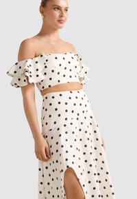 Žena v bielo odhalenom crop top s volnými ramenami a zodpovedajúcej sukni s vysokým pásom, pokrytej čiernymi bodkami a rozparkom, stojaca pred jednoduchým pozadím.
