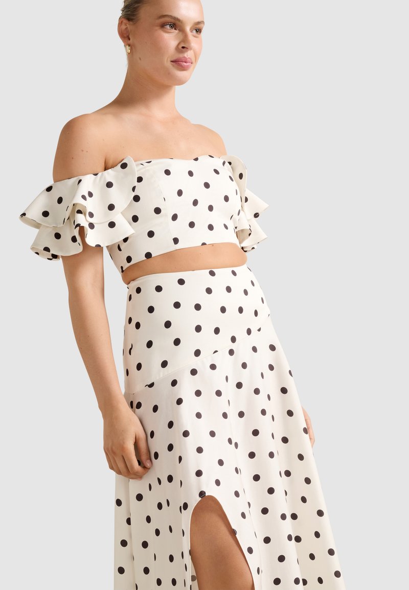 Žena v bielo odhalenom crop top s volnými ramenami a zodpovedajúcej sukni s vysokým pásom, pokrytej čiernymi bodkami a rozparkom, stojaca pred jednoduchým pozadím.